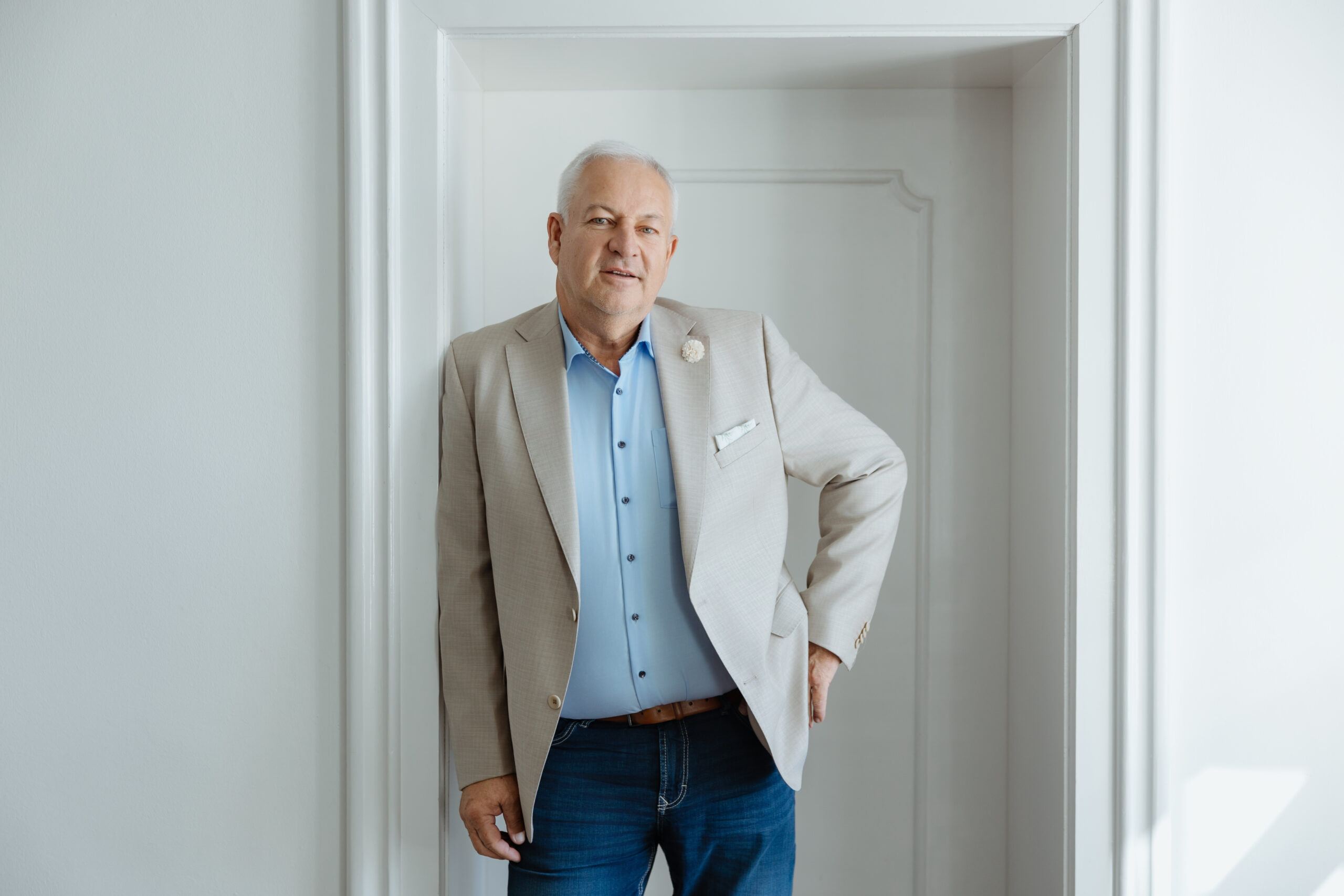 A middle-aged man in a light blazer and blue shirt poses confidently in a brightly lit interior with white walls.