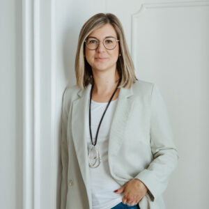 A woman with blonde hair wearing glasses and a light blazer stands confidently by a doorway, smiling softly.