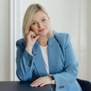 A woman in a light blue blazer sits at a dark table, resting her chin on her hand, with soft lighting in a modern setting.